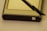 A GPS device accompanied by a stylus is positioned on a flat surface. The device has a metallic casing with a power button visible at the bottom right corner. The stylus, which appears to be black, is resting diagonally across the device.