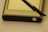 A GPS device accompanied by a stylus is positioned on a flat surface. The device has a metallic casing with a power button visible at the bottom right corner. The stylus, which appears to be black, is resting diagonally across the device.