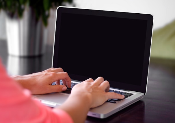 person wearing pink shirt typing on gray laptop computer on desk