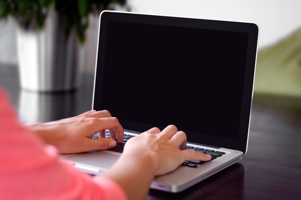 분홍색 셔츠를 입고 책상 위의 회색 노트북 컴퓨터에 타이핑하는 사람