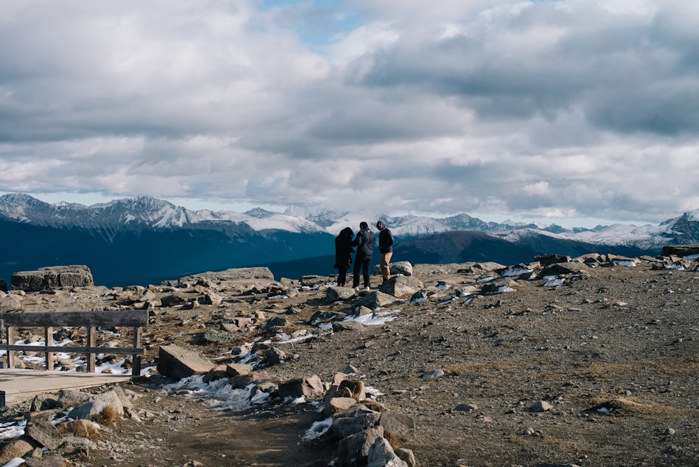 three person standing under white cloudy sky facing on snow covered mountain during daytime