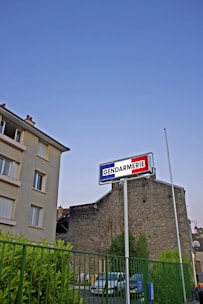 A Gendarmerie sign is prominently displayed on a tall pole against a clear blue sky. Below, there is a building complex with a stone facade, a green metal fence, and a few parked cars. The lush greenery and the calm setting suggest a residential or urban area.