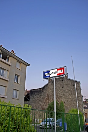 A Gendarmerie sign is prominently displayed on a tall pole against a clear blue sky. Below, there is a building complex with a stone facade, a green metal fence, and a few parked cars. The lush greenery and the calm setting suggest a residential or urban area.