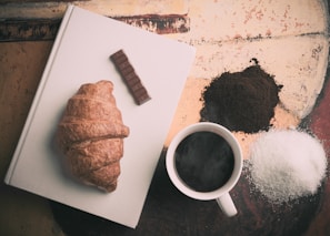 A steaming cup of coffee next to a chocolate croissant, set against a background in tones of wine red and black.