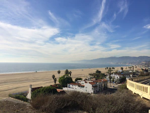 A vast sandy beach stretches alongside a calm ocean, with a clear blue sky above. Palm trees line the area near a cluster of white buildings with red roofs. In the distance, a range of mountains can be seen under the slightly cloudy sky.