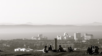 A hazy skyline with several tall buildings in the background and a group of people sitting on a grassy hill in the foreground. The distant mountains create a layered effect, adding depth to the landscape.