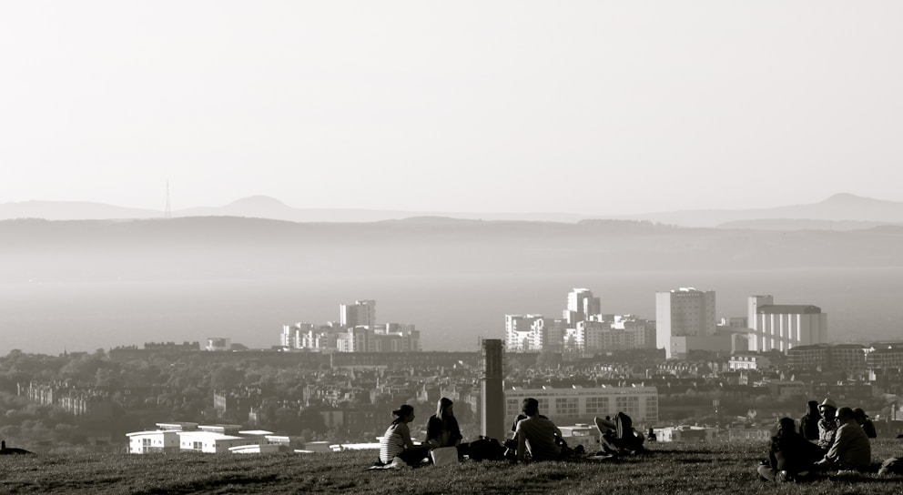 A hazy skyline with several tall buildings in the background and a group of people sitting on a grassy hill in the foreground. The distant mountains create a layered effect, adding depth to the landscape.