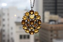 A large, circular pendant composed of multiple faceted amber-colored gemstones is suspended against a blurred urban background. The gemstones are arranged in a symmetrical pattern and set in a metal frame, creating an ornamental design.