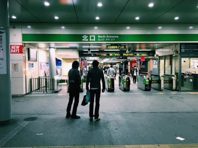 Two people stand in front of a train station entrance with turnstiles. The station is well-lit and has various signs in Japanese and English, with people visible in the background entering and exiting through the gates.