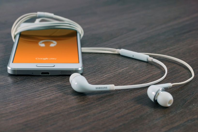 A smartphone with a visible orange screen displaying the Google Play app icon rests on a wooden surface. Earphones connected to the phone are neatly positioned beside it.