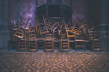A large pile of wooden chairs is stacked intricately in an indoor, dimly lit space with ornate, patterned flooring and walls. The chairs are layered on top of each other, creating a chaotic yet organized appearance.
