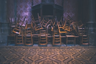 A large pile of wooden chairs is stacked intricately in an indoor, dimly lit space with ornate, patterned flooring and walls. The chairs are layered on top of each other, creating a chaotic yet organized appearance.