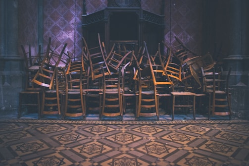 A large pile of wooden chairs is stacked intricately in an indoor, dimly lit space with ornate, patterned flooring and walls. The chairs are layered on top of each other, creating a chaotic yet organized appearance.