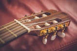 A close-up view of a classical guitar headstock displaying intricate tuning pegs and strings. The wood grain is visible, and the detail in the craftsmanship is highlighted against a soft, red background.