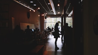 A cozy cafe interior with dim lighting and people interacting. Silhouettes of individuals are seen engaging in conversations and using laptops. Small lights hang from the ceiling, and the atmosphere is warm and inviting. Large windows in the background let in natural light, illuminating trees outside.
