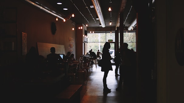 A cozy cafe interior with dim lighting and people interacting. Silhouettes of individuals are seen engaging in conversations and using laptops. Small lights hang from the ceiling, and the atmosphere is warm and inviting. Large windows in the background let in natural light, illuminating trees outside.