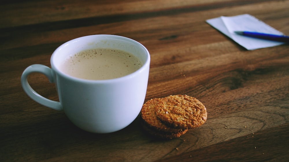 Photographie de mise au point peu profonde d’une tasse en céramique blanche à côté de deux biscuits cuits au four sur une planche en bois brun