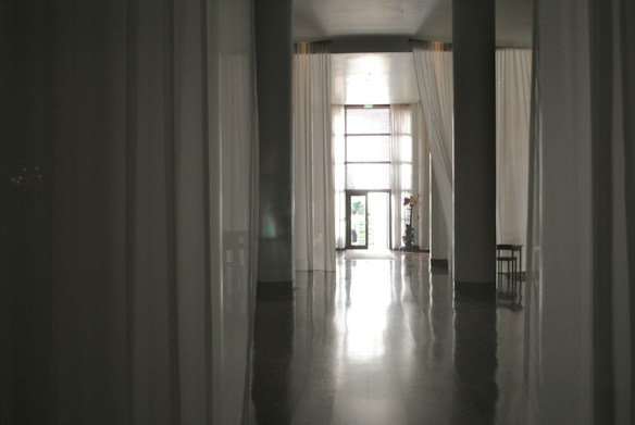 A dimly lit corridor features long, flowing curtains along the walls, leading to a large window letting in natural light. A small plant and a chair can be seen near the window, creating a serene and minimalist atmosphere.