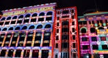 A vibrant nighttime outdoor concert illuminated by colorful 3D projection mapping on surrounding buildings.
