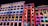 A series of buildings illuminated at night with vibrant, colorful light projections. The projections feature intricate patterns, stars, and cartoon-like characters, creating a lively and playful urban art display. The bright colors contribute to a festive atmosphere.