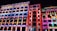 A series of buildings illuminated at night with vibrant, colorful light projections. The projections feature intricate patterns, stars, and cartoon-like characters, creating a lively and playful urban art display. The bright colors contribute to a festive atmosphere.