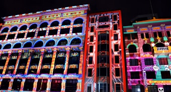 A series of buildings illuminated at night with vibrant, colorful light projections. The projections feature intricate patterns, stars, and cartoon-like characters, creating a lively and playful urban art display. The bright colors contribute to a festive atmosphere.