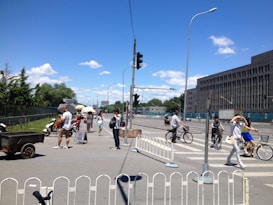 A busy urban intersection with various people crossing the street, some on foot and others on bicycles. A few individuals carry umbrellas to shield from the sun. Cars and motorcycles are also visible, and the background features a large building and greenery under a bright blue sky with scattered clouds.