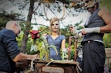 Three individuals are engaged in a theatrical scene outdoors, wearing costumes with antlered masks. One person holds a large, artificial flower, while another appears to adjust something on a wooden structure labeled 'observatoire.' The atmosphere is fantastical and surreal, set against a background of trees.