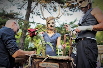 Three individuals are engaged in a theatrical scene outdoors, wearing costumes with antlered masks. One person holds a large, artificial flower, while another appears to adjust something on a wooden structure labeled 'observatoire.' The atmosphere is fantastical and surreal, set against a background of trees.