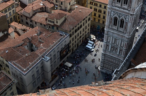 An aerial view of a busy urban scene with a cluster of historic buildings featuring reddish-brown tiled roofs. A large ornate tower with intricate designs stands prominently, with people gathered around its base. The cobblestone streets and bustling crowd suggest a popular tourist location.