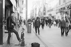 A street musician plays a guitar and sings into a microphone on a bustling city street. Passersby, some walking in pairs or groups, are visible in the background. The scene is captured in black and white, adding a timeless quality to the urban setting.