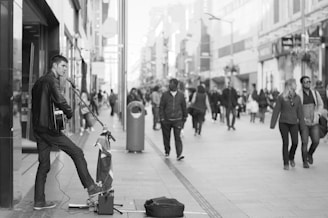 A street musician plays a guitar and sings into a microphone on a bustling city street. Passersby, some walking in pairs or groups, are visible in the background. The scene is captured in black and white, adding a timeless quality to the urban setting.