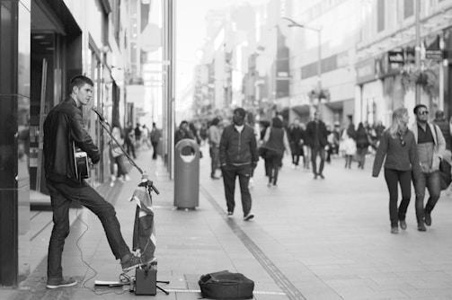 A street musician plays a guitar and sings into a microphone on a bustling city street. Passersby, some walking in pairs or groups, are visible in the background. The scene is captured in black and white, adding a timeless quality to the urban setting.