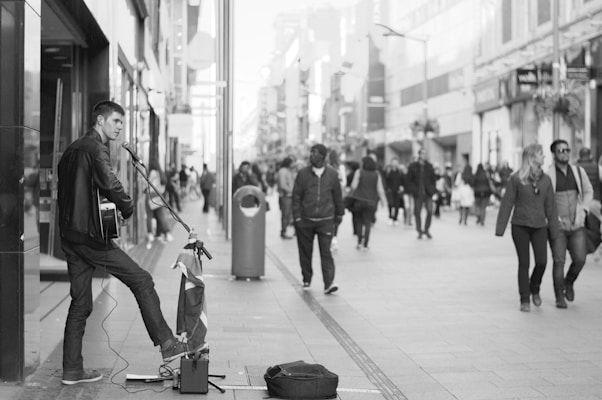 A street musician plays a guitar and sings into a microphone on a bustling city street. Passersby, some walking in pairs or groups, are visible in the background. The scene is captured in black and white, adding a timeless quality to the urban setting.