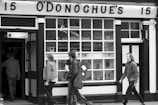 Black and white photograph of people walking past a pub named O'Donoghue's. A person is carrying a guitar case. The facade has large windows and a sign with the number 15.