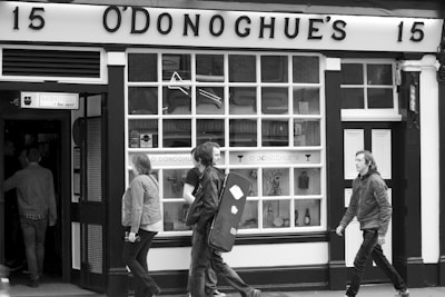 Black and white photograph of people walking past a pub named O'Donoghue's. A person is carrying a guitar case. The facade has large windows and a sign with the number 15.