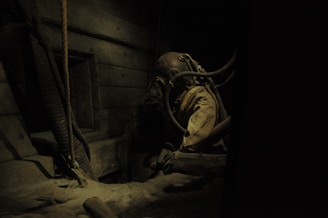 In a dimly lit and confined space, an individual is wearing a vintage diving suit complete with a helmet and hoses. The setting is suggestive of an old, possibly wooden ship or underwater environment, with visible ropes and pipes surrounding the figure.