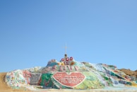 A colorful painted hill with religious messages and a central cross on top. The artwork includes large red letters reading 'God is Love', surrounded by a heart with inscriptions. Various colors like green, blue, yellow, and red are used in different patterns and messages across the hill.