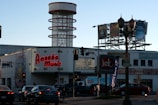 A busy urban intersection features a large building with a neon sign for Amoeba Music, advertising CDs, LPs, and DVDs, and offering to buy, sell, or trade. In the background is a cylindrical structure with red rings. Several cars are stopped at the traffic lights in the foreground, and there are billboards visible on top of the building promoting different music albums. A Jack in the Box fast-food restaurant sign is also prominent in the scene.