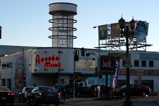 A busy urban intersection features a large building with a neon sign for Amoeba Music, advertising CDs, LPs, and DVDs, and offering to buy, sell, or trade. In the background is a cylindrical structure with red rings. Several cars are stopped at the traffic lights in the foreground, and there are billboards visible on top of the building promoting different music albums. A Jack in the Box fast-food restaurant sign is also prominent in the scene.