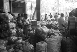 A bustling market scene with several people engaged in various activities. Large sacks and bundles of produce are stacked high, including what appears to be onions, coconuts, and other vegetables. Most of the individuals in the scene are dressed in casual clothing, and the setting has an open, shaded area with more produce visible in the background.