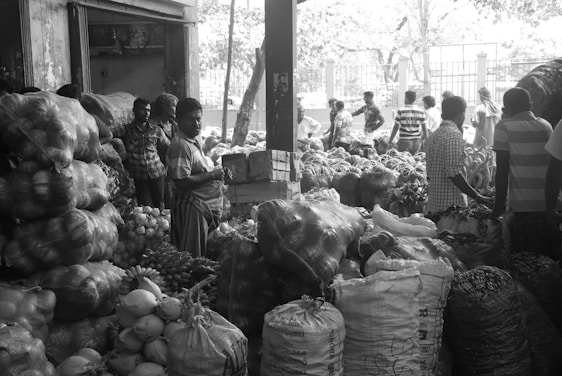 A bustling market scene with several people engaged in various activities. Large sacks and bundles of produce are stacked high, including what appears to be onions, coconuts, and other vegetables. Most of the individuals in the scene are dressed in casual clothing, and the setting has an open, shaded area with more produce visible in the background.
