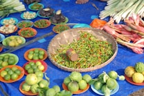A vibrant market display featuring an assortment of fresh produce arranged on colorful plates. In the center, a woven basket is filled with red and green chili peppers, with other plates surrounding it containing limes, lemons, eggplants, and other vegetables. Stalks of lemongrass and ginger flowers are neatly bundled nearby, all set on a blue tarp.