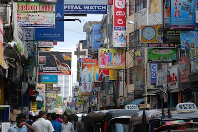 A bustling street filled with colorful storefronts and numerous signs in different languages. The area is crowded with people walking and vehicles lined up, some of which are tuk-tuks with 'Meter Taxi' written on them. Clothing advertisements and phone store signs are prominently displayed.