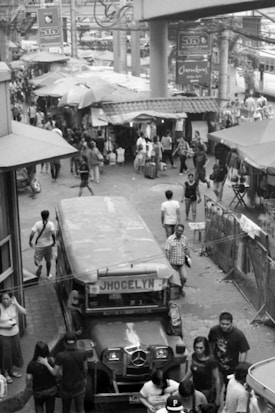 A bustling street scene in black and white, featuring a jeepney with a sign 'JHOCELYN' at the forefront. The street is crowded with people walking in various directions. Numerous market stalls are visible with umbrellas providing shade. Overhead, there are multiple signs and an elevated structure, possibly part of a transport system.