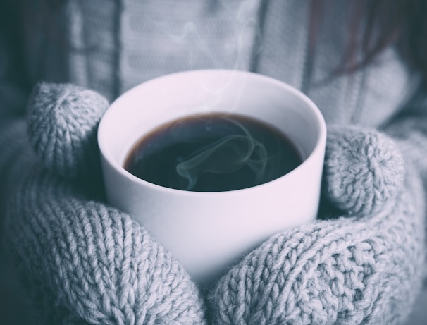 A close-up of warm hands wrapped around a steaming mug near a heater.