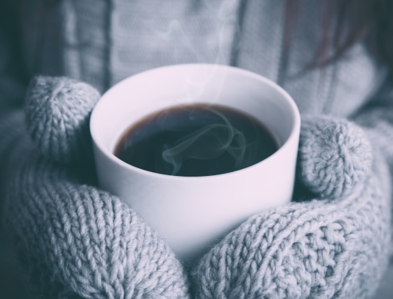 A close-up of hands wearing soft, knitted gloves holding a steaming cup of cocoa on a snowy day.