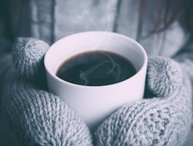 A cozy scene featuring a pair of hands in thick, gray knitted mittens holding a steaming cup of coffee or tea. The photograph emphasizes warmth and comfort during a chilly day.