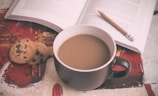 A cozy scene with a steaming cup of coffee in a dark mug placed on a surface with abstract patterns. Nearby are two chocolate chip cookies placed beside an open book. A pencil rests between the pages, suggesting a moment of relaxation and thought.