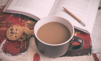 A cozy scene with a steaming cup of coffee in a dark mug placed on a surface with abstract patterns. Nearby are two chocolate chip cookies placed beside an open book. A pencil rests between the pages, suggesting a moment of relaxation and thought.